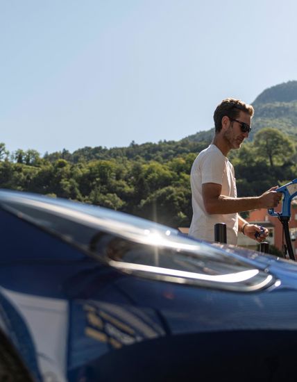Man charging an electric car at a modern station with green hills in the background.