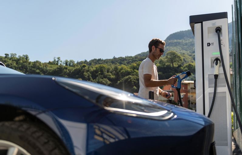 Uomo ricarica un'auto elettrica presso una colonnina, con colline verdi sullo sfondo.