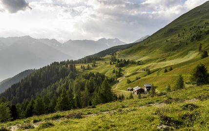 Sonnige Almlandschaft mit verstreuten Hütten, Wäldern und Bergkulisse unter teils bewölktem Himmel.